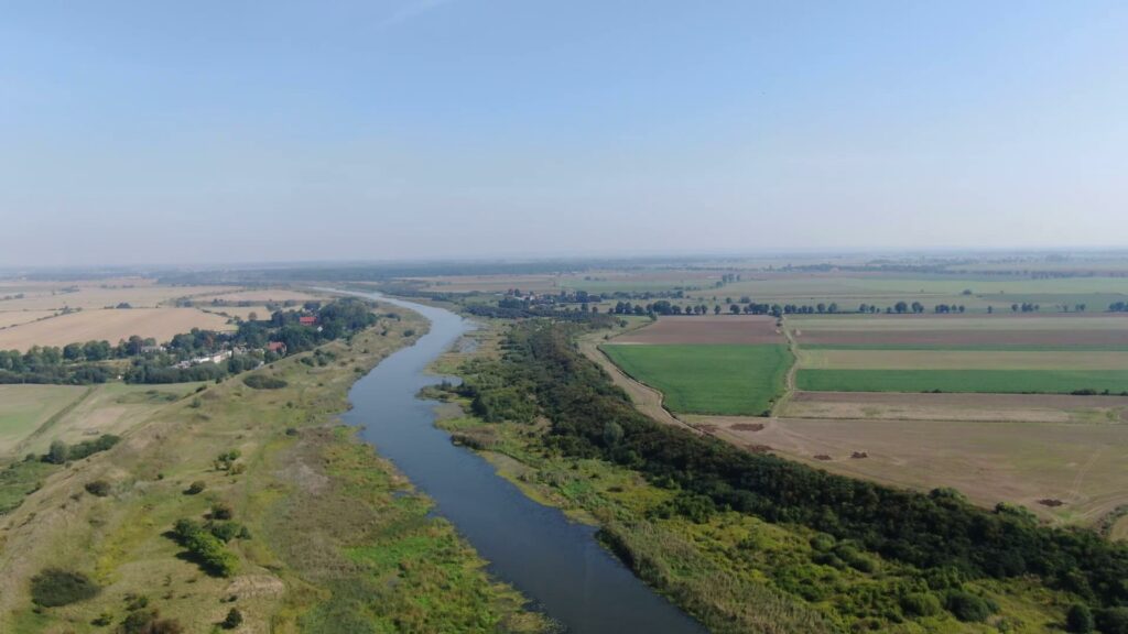 Krajobraz z lotu ptaka. Środkiem płynie rzeka po obu jej stronach biegnie wał przeciwpowodziowy. W oddali widać pola uprawne drzewa i pojedyncze zabudowania.